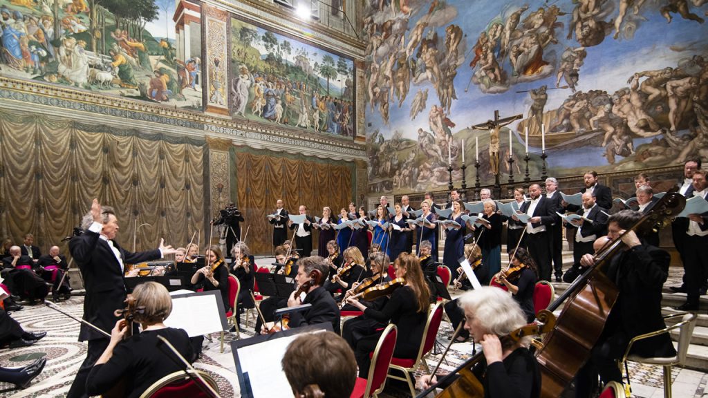 Harry Christophers conducting The Sixteen choir inside the Sistine Chapel. In front of the choir is a seated orchestra.