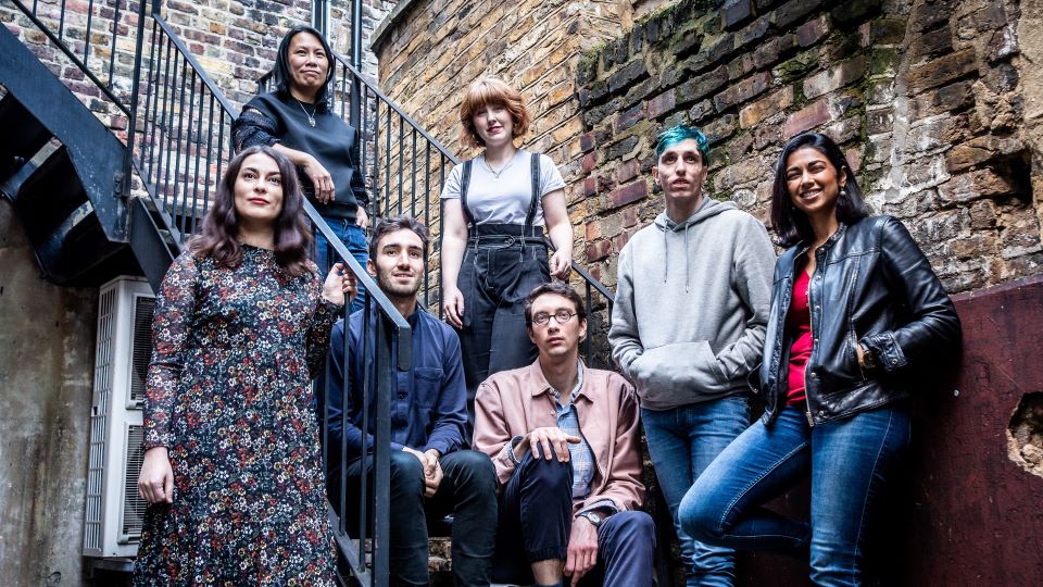 A photo of the 2020 to 2021 Genesis Almeida Playwrights cohort. The playwrights are stood along a metal staircase outside a building with brown brick walls.