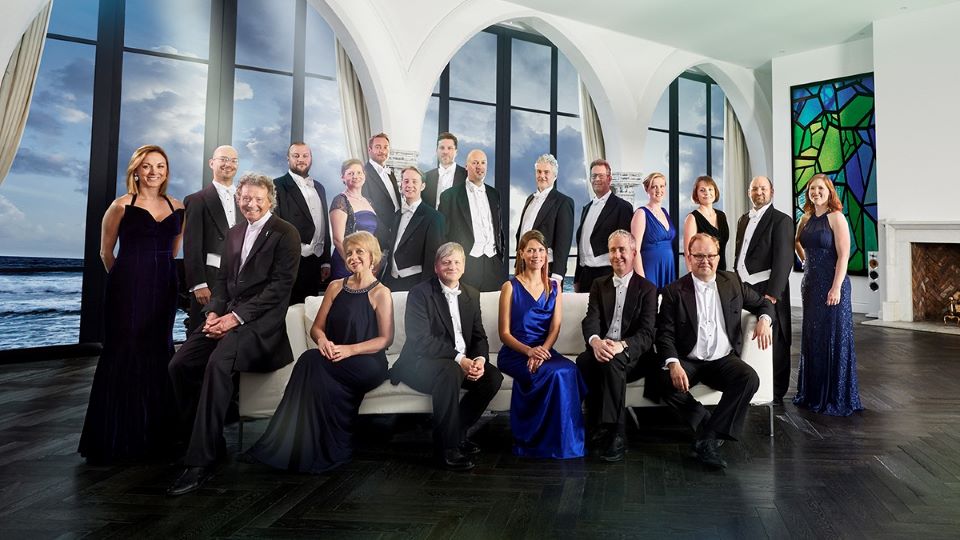 A photo of Harry Christophers and The Sixteen. The group are inside a large bright room with 3 arched windows at the back looking out onto a blue sky. The group are arranged across a white sofa, either sitting on it or standing behind it. The group are all dressed in smart black suits or long rich blue dresses. Everyone is smiling at the camera.