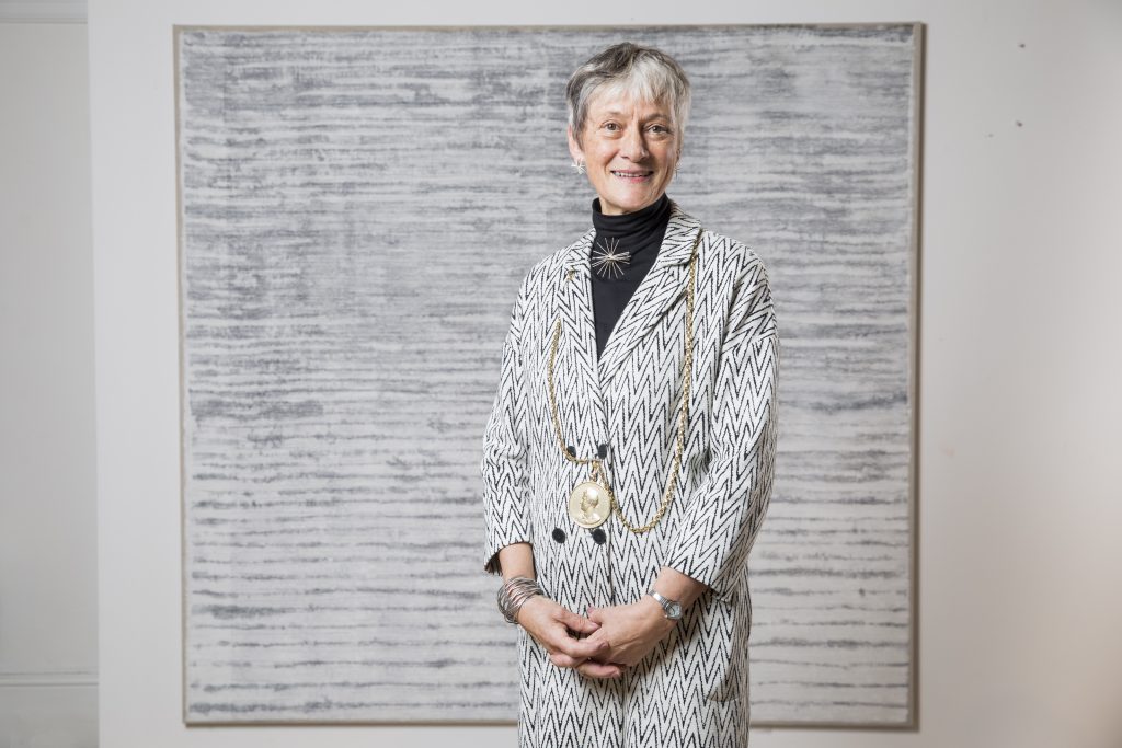 A photo of Rebecca Salter stood in front of a black and white art work, wearing a similarly coloured jacket with a black top underneath and a long gold necklace. Rebecca has short grey hair, and is holding her hands together in front of her and smiling at the camera.