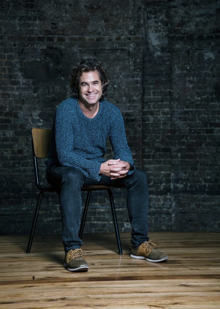 A photo of Rupert Goold, the Artistic Director of the Almeida Theatre. Rupert is sat on a chair inside a room with a wooden floor and dark grey brick walls. He has medium length curly hair, is wearing a woollen jumper and jeans, smiling at the camera.