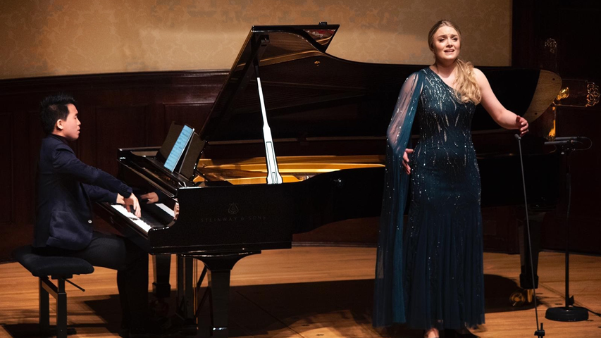 A photo of Camilla Harris, who is from Genesis Sixteen cohort 1, singing onstage with a pianist. Camilla has long blonde hair which she is wearing in a side ponytail that comes down in front of one shoulder. She is wearing a long blue dress that has one long mesh sleeve. The pianist is to the left of Camilla, and is wearing a navy blue suit while they play the piano.