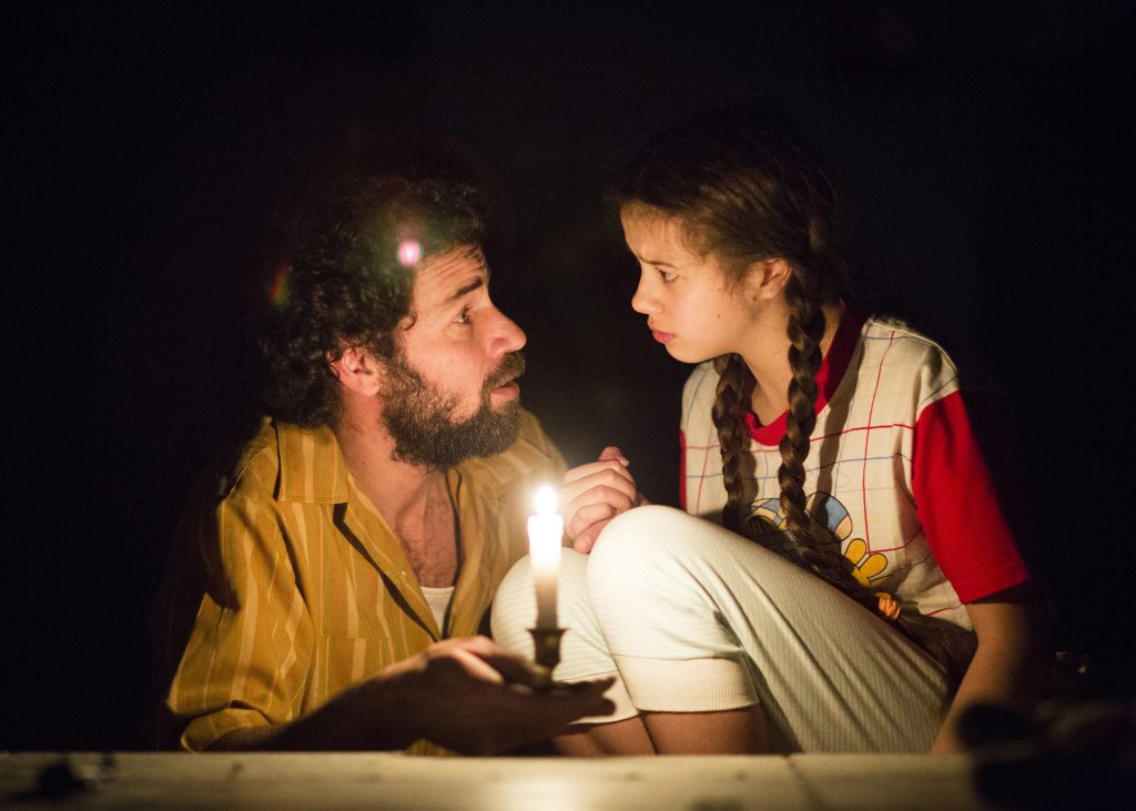 A photo of two actors in the middle of a scene. The actor on the left is a man in his thirties with short dark hair and a dark beard. He is holding a candle in front of him. The actor on the right is a young girl aged between 6 and 10, with long brown hair in two plaits. She is crouched down, looking directly at the man.