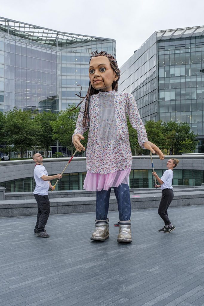A photo from Good Chance Theatre's 'The Walk' project. The photo is of a hug puppet which is 3 times the size of the two people operating it. The puppet is of a little girl with long brown hair in a patterned shirt, a pink skirt, blue leggings, and silver boots. The puppet and two people operating it are outside on the London Southbank.