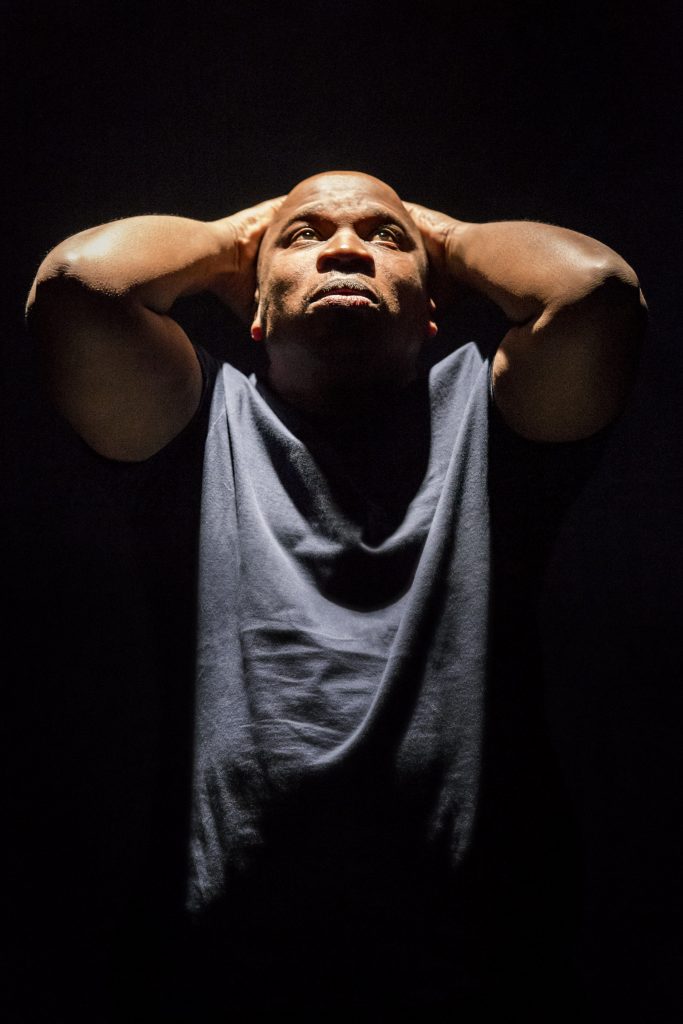 A photo of an actor onstage lit by a bright spotlight. The actor has a shaved head and is wearing a blue t-shirt. They are holding both hands on the top of their head, tilting their head and looking upwards.