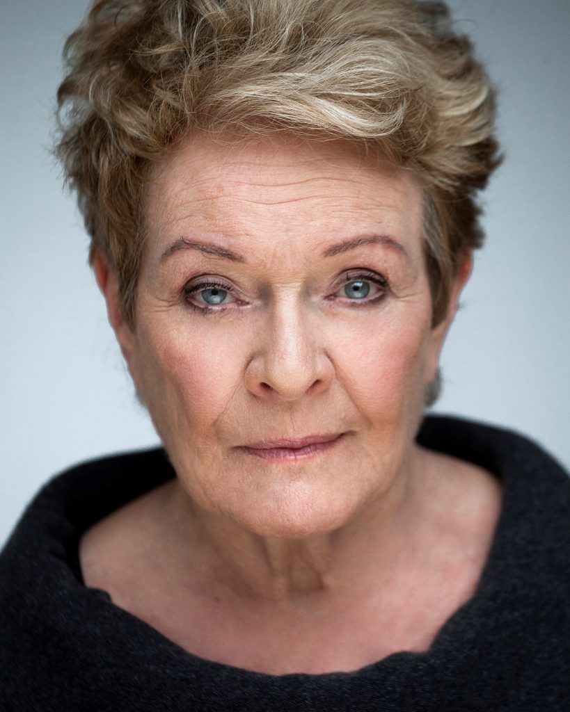 A headshot of Dame Janet Suzman DBE, in front of a plane grey background. Janet has short blonde hair and is wearing a black top with a wide neck. She is looking at the camera with a neutral expression.