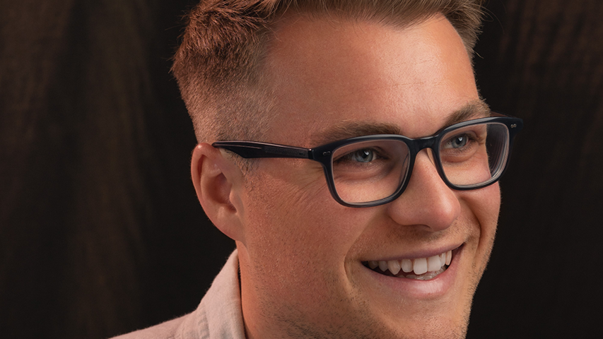 A headshot of singer Jonathan Hanley, who is from the Genesis Sixteen cohort 6. Jonathan has short blonde hair and is wearing glasses with a black frame and a white shirt. He is looking to the right side of the shot and is smiling at something off camera. Behind him is a plane black background.