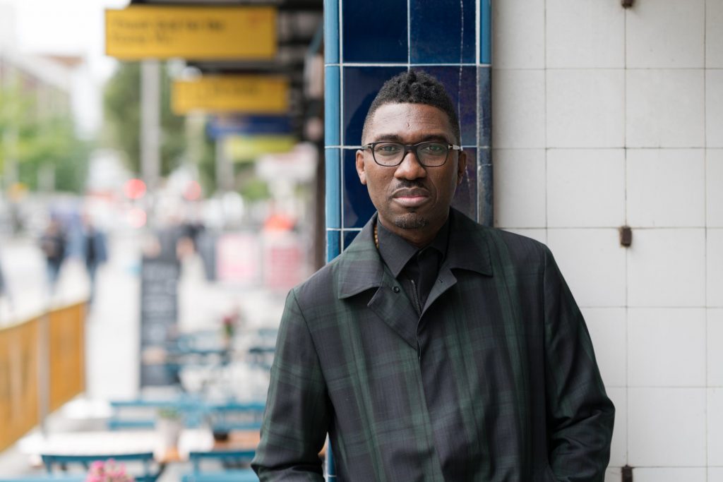 A photo of Kwame Kwei-Armah stood outside against a blue tiled wall. He has short dark hair, and is wearing glasses, and a dark green and black tartan coat. He has a neutral expression, and is looking directly at the camera.