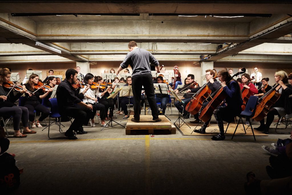A photo of The Multi-Story Orchestra. The photo is taken inside an indoor car park with stone walls and ceiling. In the centre is a conductor who is stood on a wooden podium with their arms in the air in front of them. They are facing away from the camera. Around the conductor is a large seated orchestra playing violins and cellos.