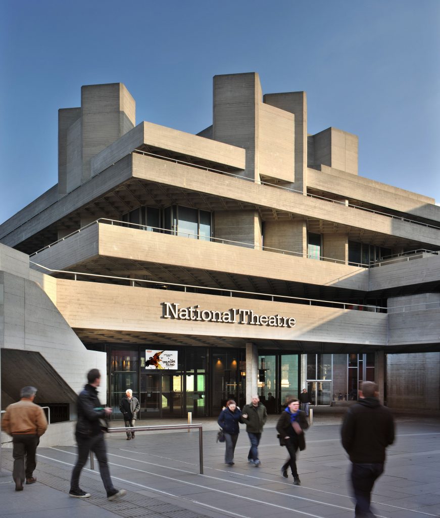 A photo of the outside of the National Theatre building. The building has layered floors which have a light stone exterior. The layers cross over each other, interlocking so it looks like a Chinese puzzle. Across the front of the building, above an automatic door is the text 'National Theatre'. In front of the building are crows of pedestrians walking by.