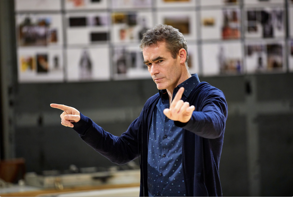 A photo of Rufus Norris. He is a middle aged man with short grey hair. He is wearing a dark blue shirt and cardigan, and is holding his hands out either side of him, as if directing someone off camera.