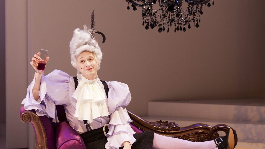 A photo of actor Samuel Barnett performing at the Sheffield Crucible Theatre. Samuel is dressed in a period Georgian men's costume and tall grey wig. He is laying across a chaise long, and is holding up a glass of wine as if cheersing. Behind him are some plain white steps and a black chandelier. 