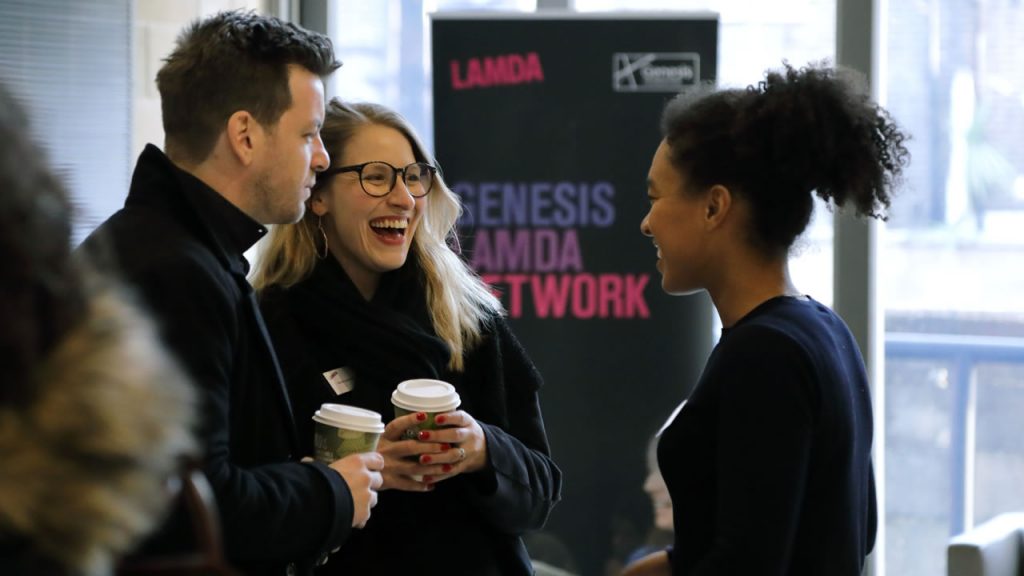 A photo of two people inside a bright foyer with a window behind them and a large stand-up board with the text 'Genesis LAMDA Network' and the 'LAMDA' and 'Genesis Foundation' logos. The two people are chatting and laughing with each other. One of the people is holding a takeaway cup of coffee.
