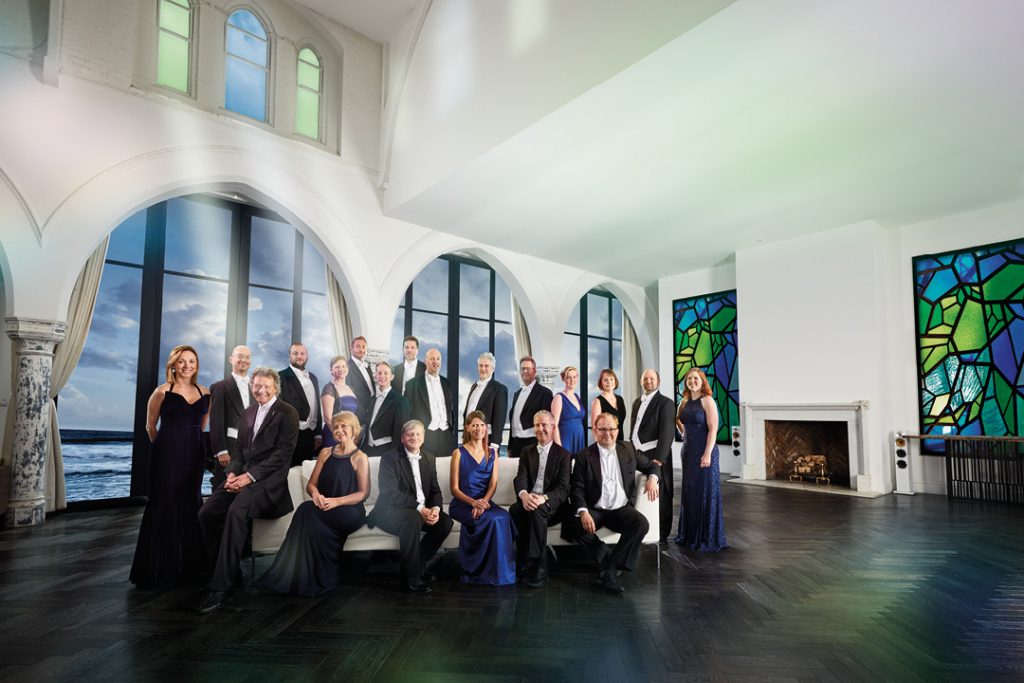 A photo of The Sixteen in a grand looking room, dressed in smart attire, smiling at the camera. Some members are stood up behind a sofa, and some members are sat down on the sofa.