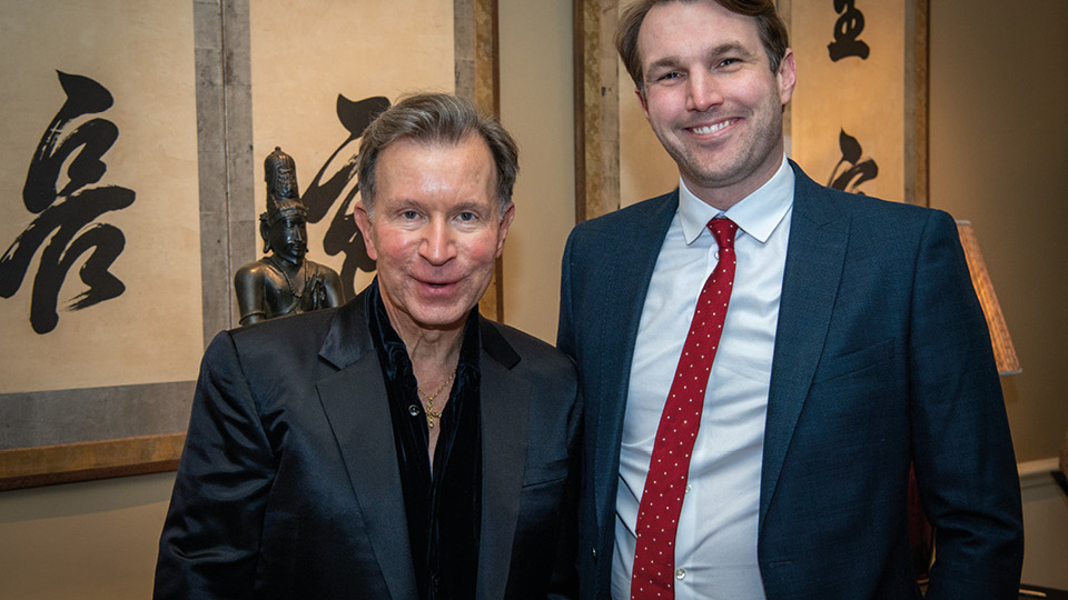 John Studzinski and George Turvey stand side-by-side smiling, dressed in smart attire. They are standing in a decorated hallway with paintings and a small statue.