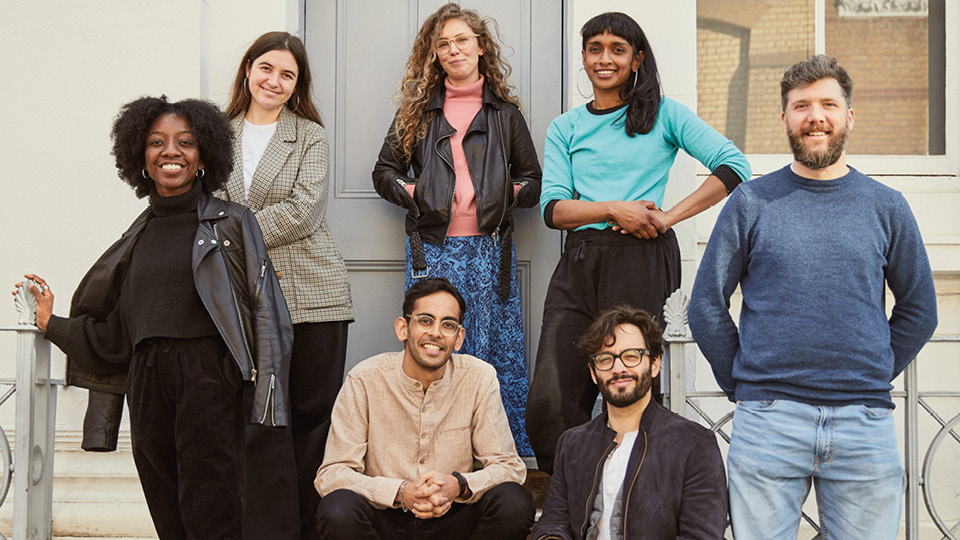A photo of the current cohort of the Genesis Almeida New Playwrights, Big Plays Programme. The seven playwrights are stood outside on steps in front of a white building, smiling at the camera.