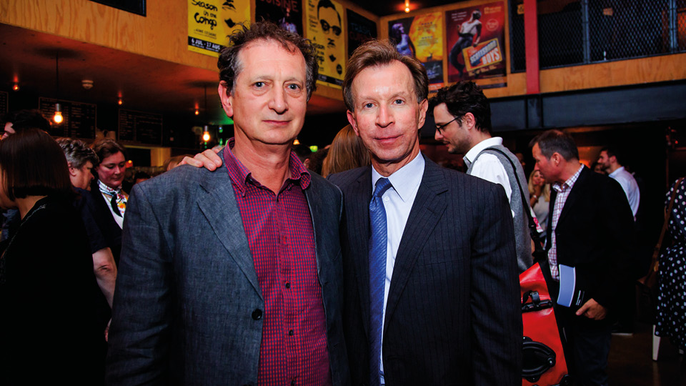 A photo of David Lan (left) and John Studzinski (right). David is a white man with short hair, wearing a burgundy shirt and a grey suit jacket. John has his arm around David. John is a white man with short hair, wearing a white shirt, blue tie, and navy suit jacket. They are stood in a crowded room.