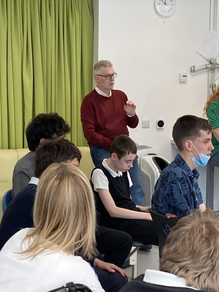 In a large classroom, James Macmillan is standing behind several students who are sat down. James has short grey hair, glasses, and is wearing a burgundy jumper with a white shirt underneath. He is conducting with one hand.