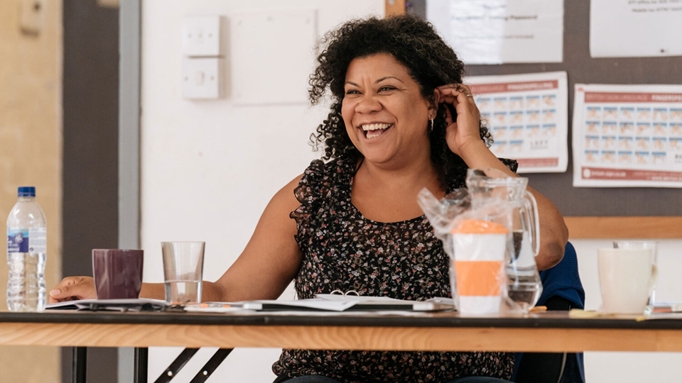 A landscape photo of Nancy Medina sat at a desk. Nancy has brown medium length with tight curls. She is wearing a black top with light colour flowers on, and is smiling off camera. She is sat inside a rehearsal room with scripts, a glass and a reusable coffee cup on the desk.