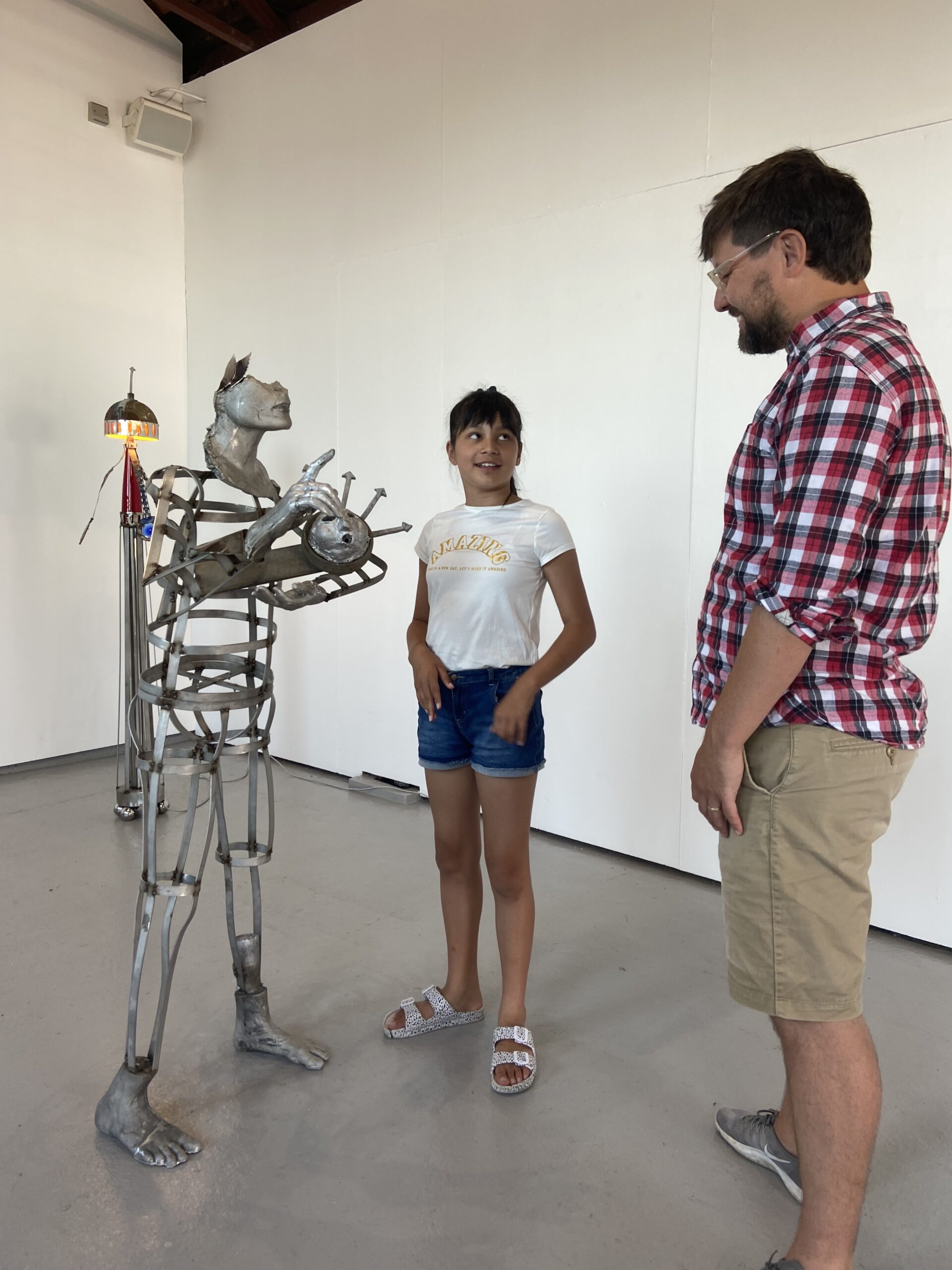 A metal sculpture of a person with half a face inside an art studio. Stood next to it is a a child in white shirt and denim shorts, smiling at an adult who is looking at the sculpture, wearing a checked shirt and brown shorts. 