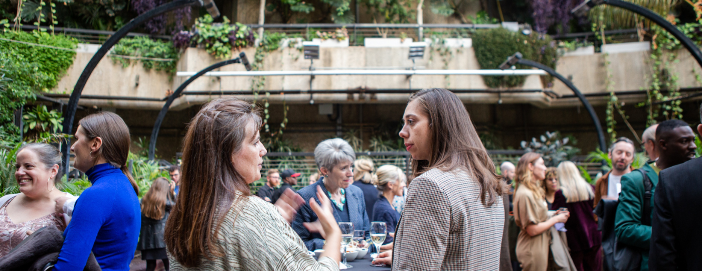 A wide shot of inside the garden at the Barbican Centre. The walls are a mixture of brick and leaves, with branches and foliages hanging from every direction. On the ground is busy with people dressed in business attire, having drinks and talking to one another.