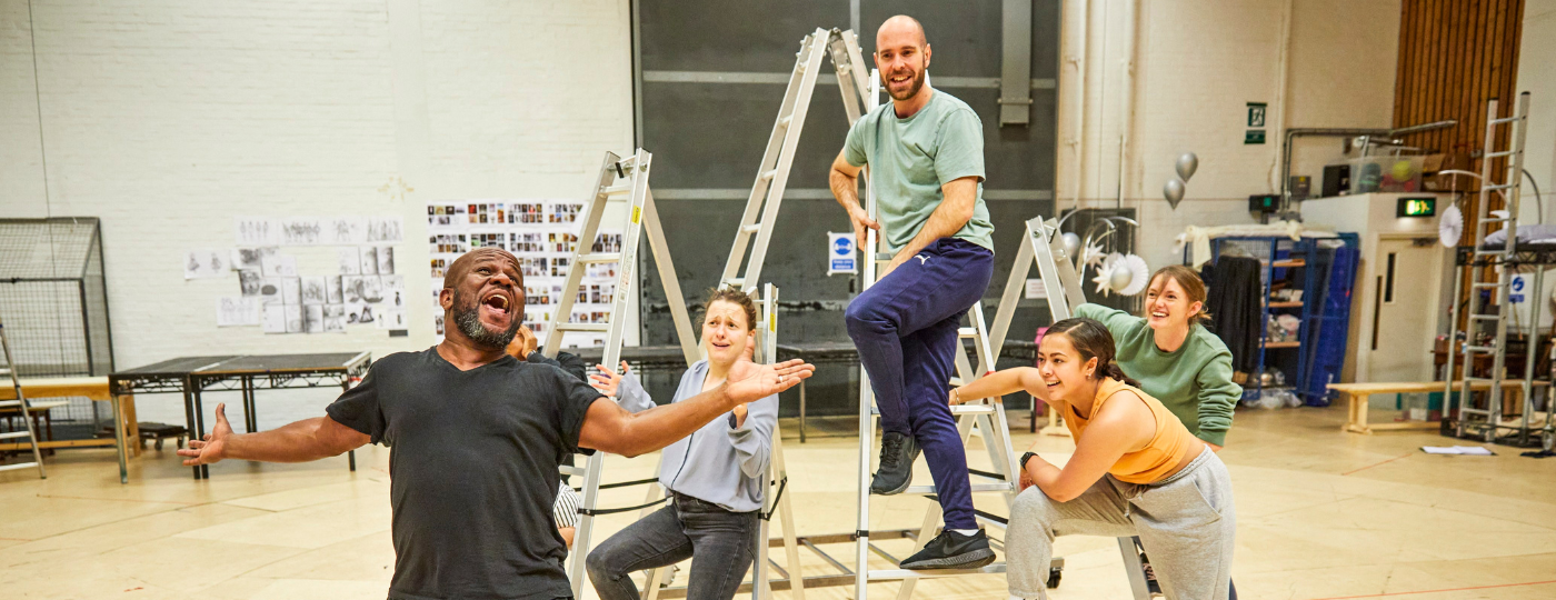 Inside a large rehearsal room with white brick walls and a wooden floor, a group of four actors stand on 3 different step ladders, smiling and laughing at another actor who is stood in the foreground of the photo, with their arms spread out wide, laughing with a big open smile.