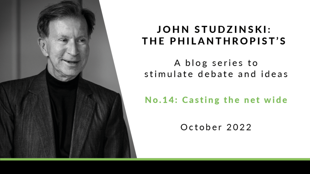 The left side of the image is a black and white photo of John Studzinski in a dark grey suit and black top, smiling off camera. The right of the image has a white background with the text 'John Studzinski: The Philanthropist's View. A blog series. stimulate debate and ideas. No. 14: Casting the net wide. October 2022.