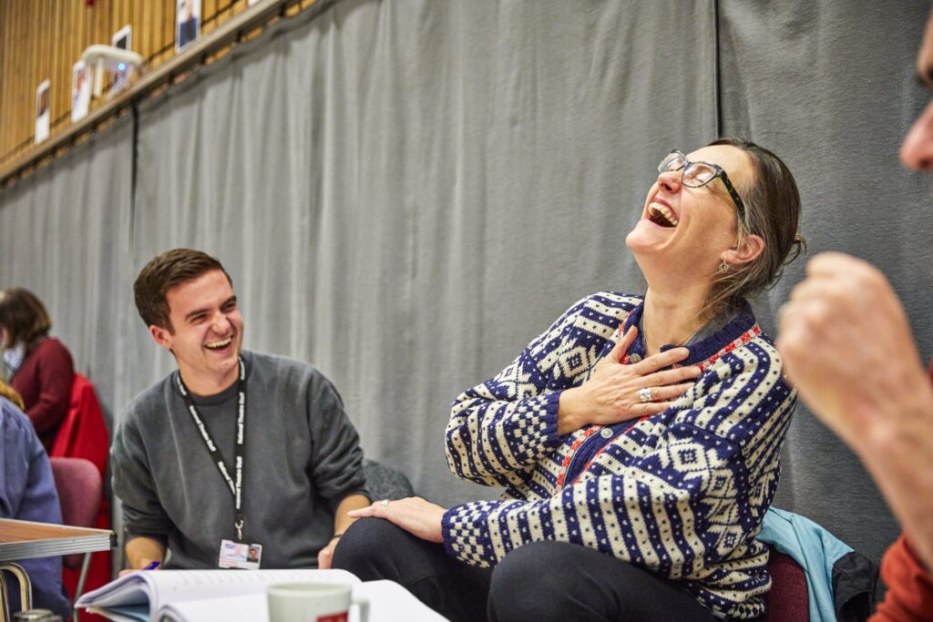 A photo of two people sat in a rehearsal room. The person on the right who is closer to the camera, is holding one hand to their chest, and throwing their head back laughing. The person on the left is sat slightly further back and is laughing with them.