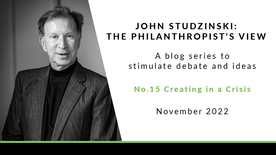 The left side of the image is a black and white photo of John Studzinski in a dark grey suit and black high-neck top, looking at the camera with a neutral expression. The right of the image has a white background with the text 'John Studzinski: The Philanthropist's View. A blog series. stimulate debate and ideas. No. 15: Creating in a Crisis. November 2022'.