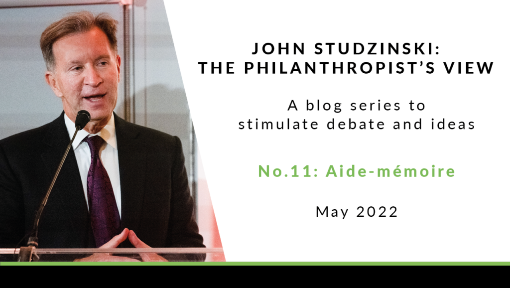 To the left is a photo of John Studzinski, stood at a microphone, wearing a black jacket, a white shirt, and a dark purple tie. He has short grey hair, and is talking, looking to the side. To the right of the image is a white background with black text saying 'John Studzinski - Philanthropist's View. A blog series to stimulate debate and ideas. No. 11: Aide-mémoire. May 2022'.