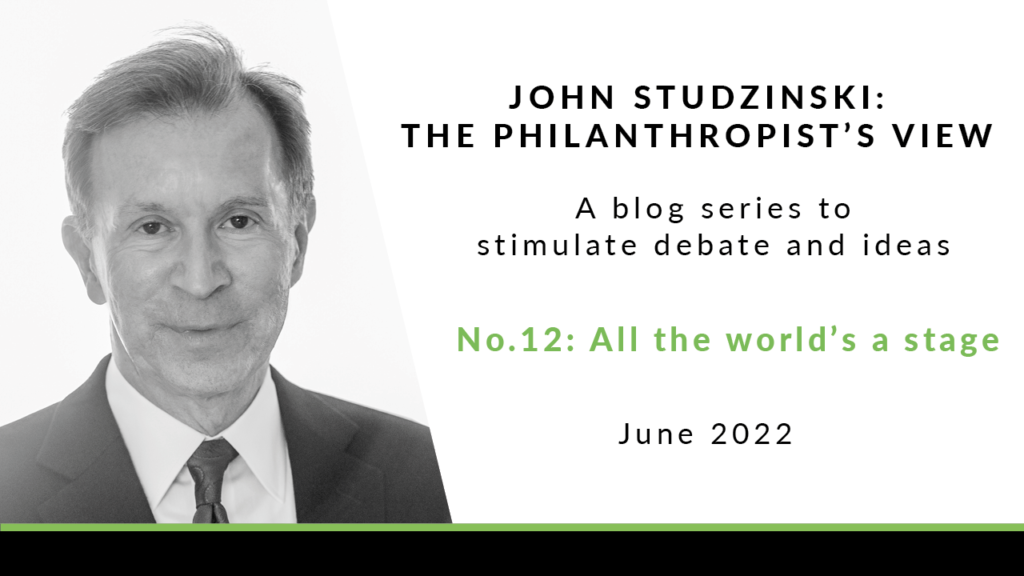 To the left is a black and white headshot of John Studzinski, wearing a black jacket, white shirt, and black tie. He has short grey hair, and is looking directly at the camera with a neutral expression. To the right of the image is a white background with black text saying 'John Studzinski - Philanthropist's View. A blog series to stimulate debate and ideas. No. 12: All the world's a stage. June 2022'.