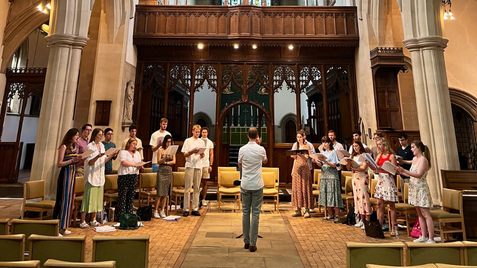 The 11th Genesis Sixteen cohort stood inside a grand looking church, holding sheets of music, singing out to empty pews. In front of them, facing away from the camera, is a conductor.