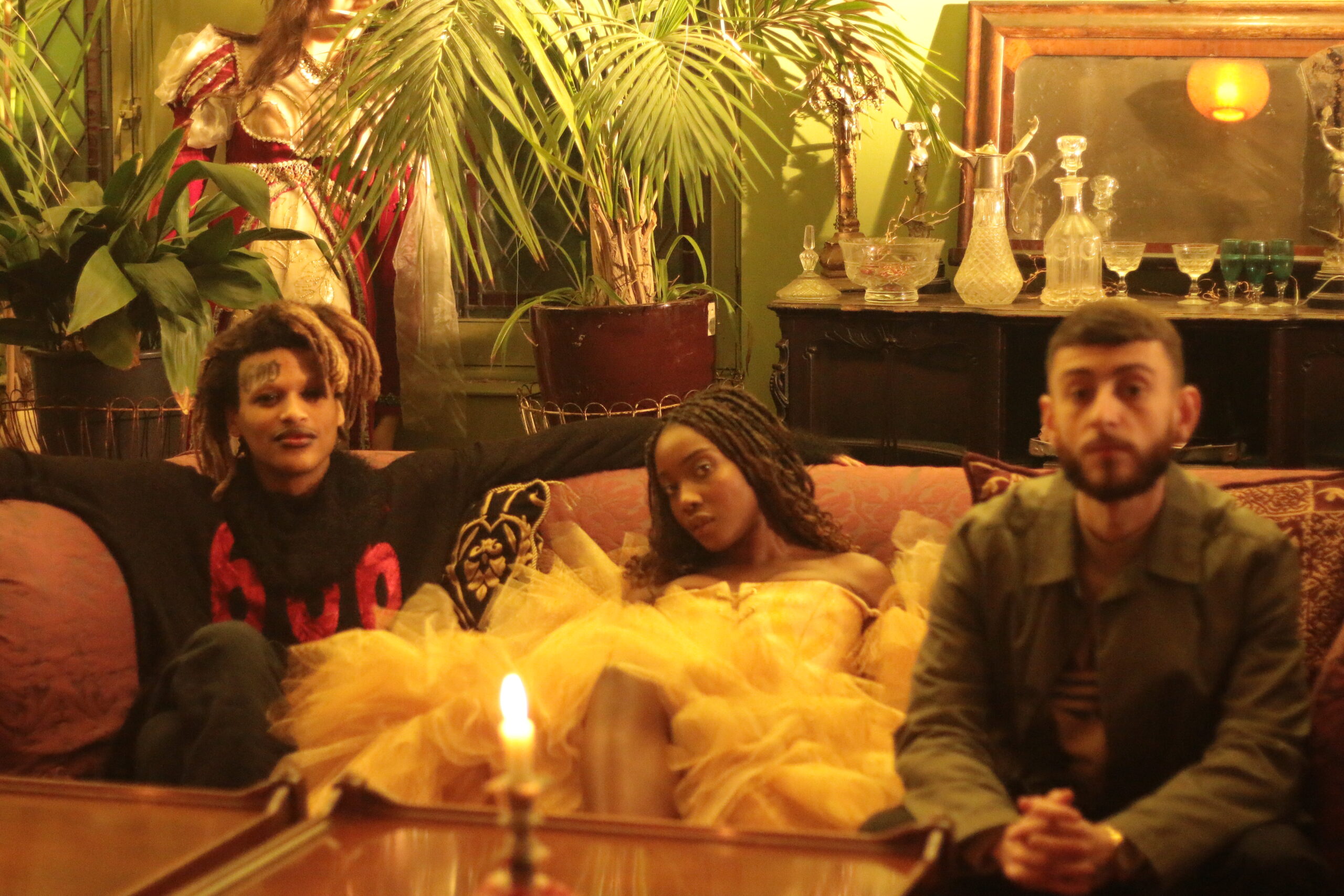 Inside a dimly lit room with a tall green plant at the back, and several glass ornaments sitting on a wooden cabinet. Three people sit on a sofa with a glass coffee table in front of them. A single lit candle sits on the coffee table. The three people look directly at the camera with neutral expressions.