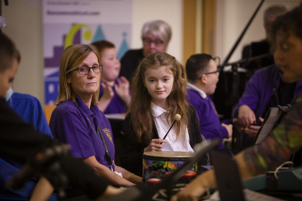 Inside a busy classroom with school pupils in school uniforms, an adult with medium length blonde hair and a purple top sits with a child with long brown hair at a table with musical instruments. The child is holding a drum stick for a drum in front of them.