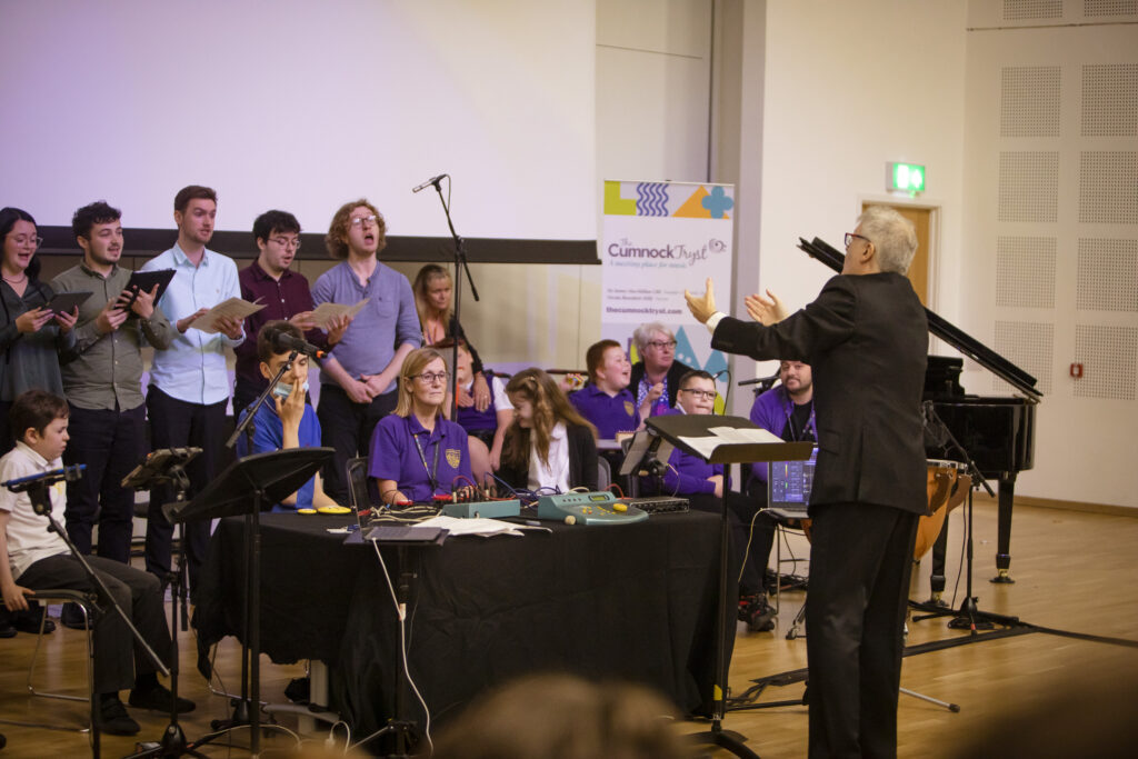 Inside a large classroom, a group of school pupils in school uniforms sit with sheets of music on a table in front of them, or stand holding the sheets of music. In front of them, facing away from the camera, is James MacMillan who is conducting.