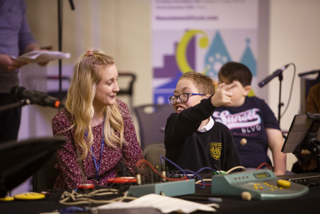 An adult with long blonde hair and a pink top sits with a child wearing glasses and a school uniform at a table with electronic musical instruments on. The child is looking at the adult, smiling and pointing forwards. 