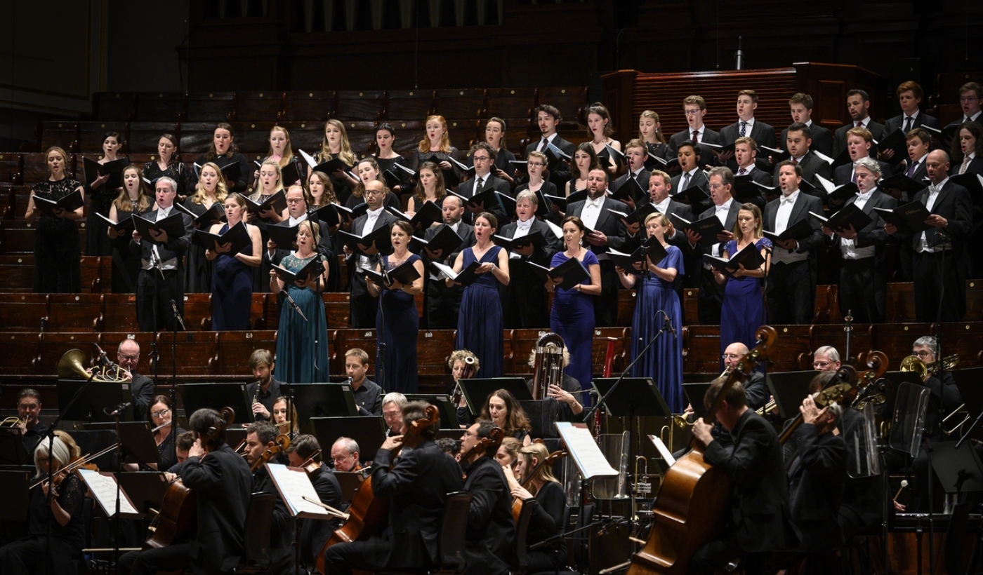 The Sixteen choir and members of Genesis Sixteen choir stand in rows, holding open song books, singing out. In front of them is a seated orchestra.