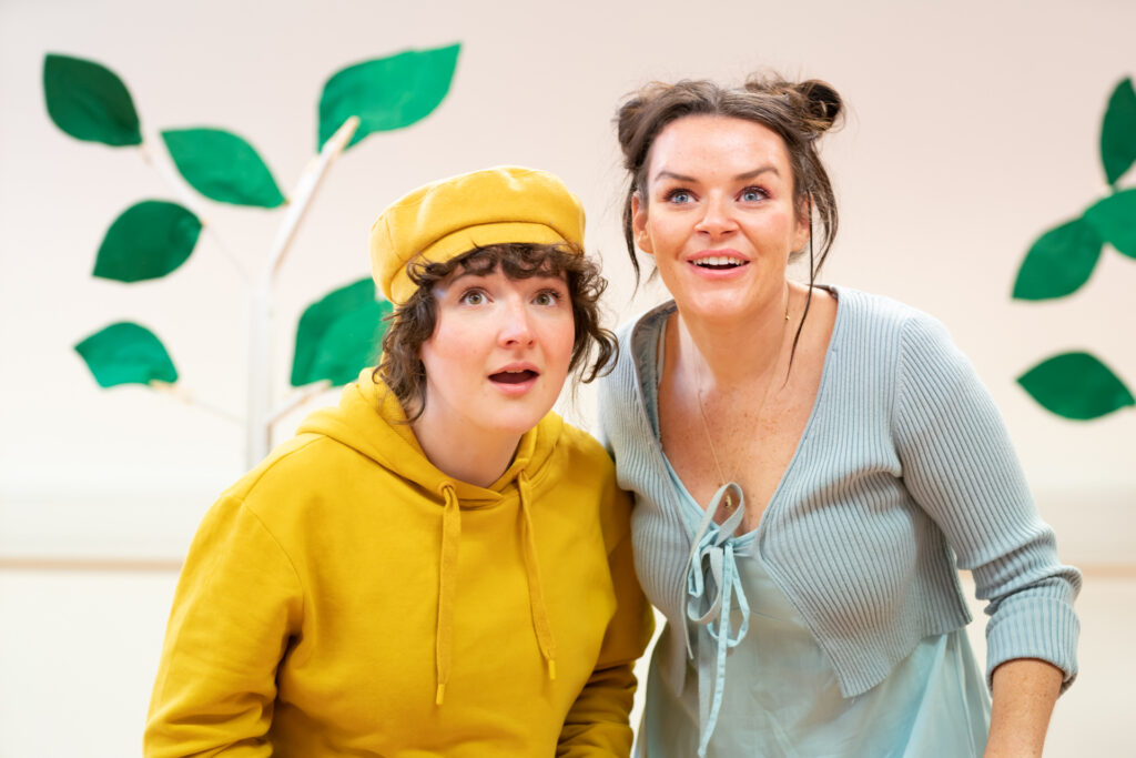 In front of a white wall and a fake tree, two actors stand beside each other, looking out into the distance, and holding on hand next to their mouth, as if calling out. The actor on the left has curly brown hair that is tucked into a mustard cap, and is wearing a matching mustard jumper. The actor on the right has brown hair tied up into two high buns. and is wearing a light blue dress with a matching cardigan.