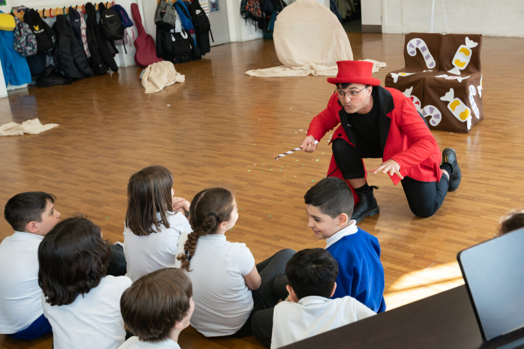Inside a large school hall, an actor wearing a red top hat and waistcoat holds a magic wand, and crouches in front of a group of smiling school children.