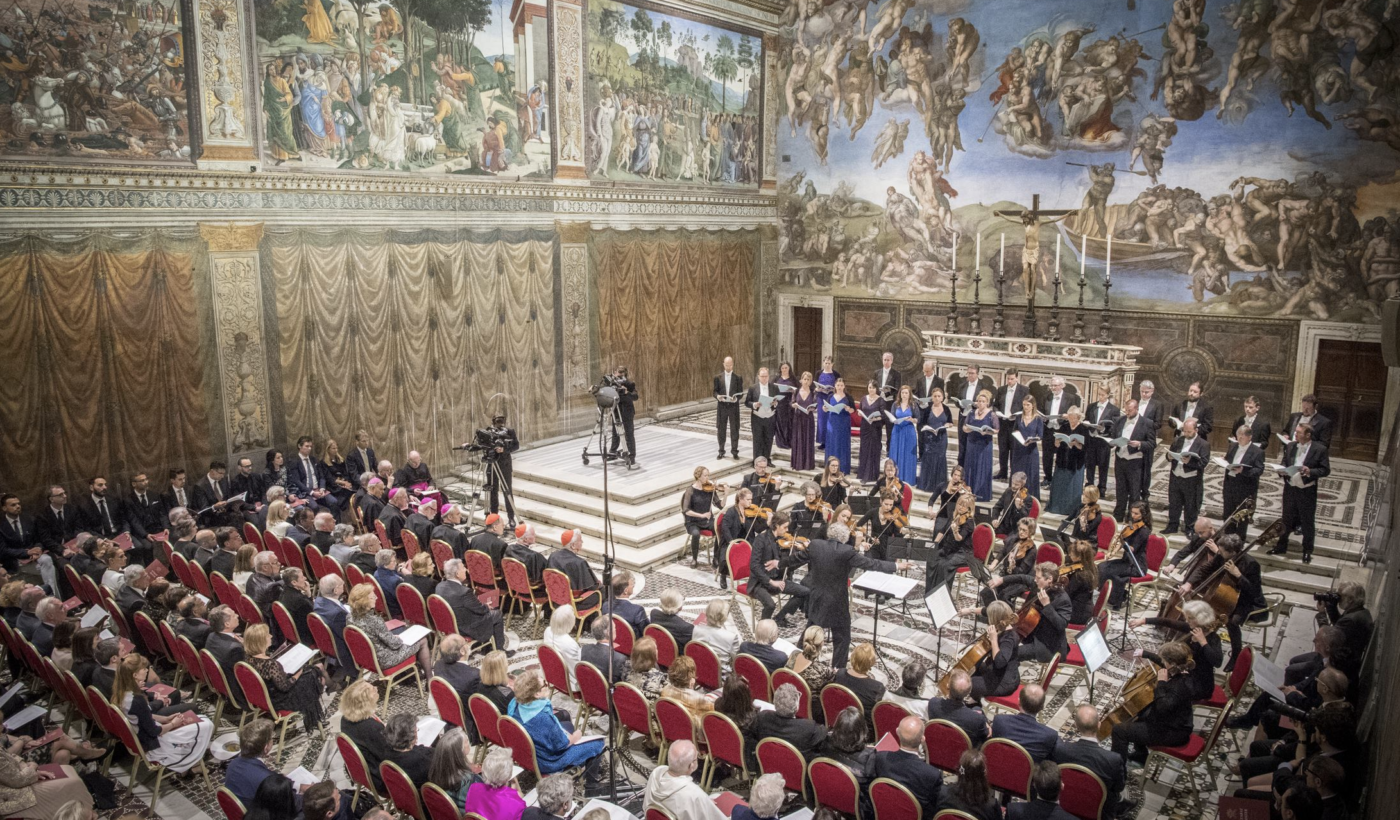 A wide shot of The Sixteen choir performing in front of a large audience inside the Sistine Chapel. The walls of the chapel have large grand paintings which take up the entire wall.