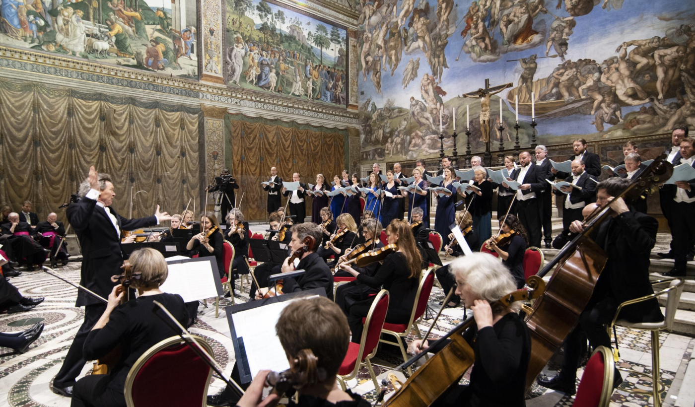 Harry Christophers inside the Sistine chapel, conducting The Sixteen choir, in front of an orchestra.