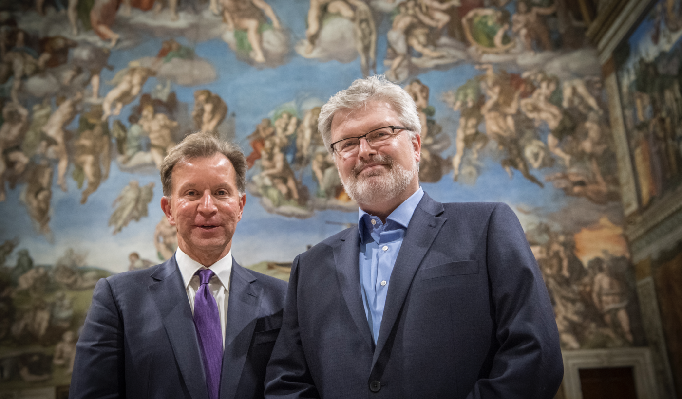 John Studzinski & Sir James MacMillan dressed in suits, stood in front of a large old painting, smiling at the camera.