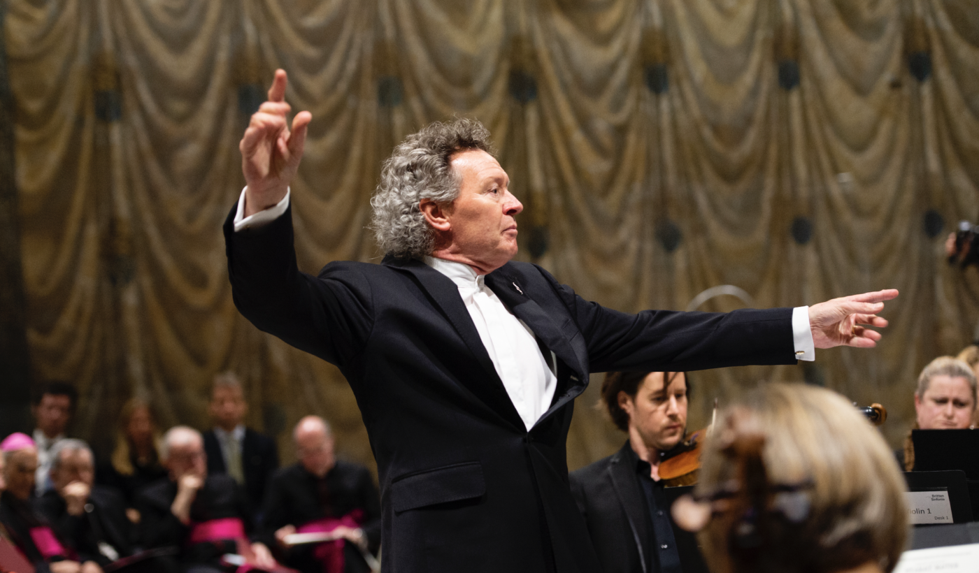 Inside the Sistine Chapel. A close up of Harry Christophers dressed in a black suit and white shirt, conducting an orchestra with his arms held dramatically in the air.