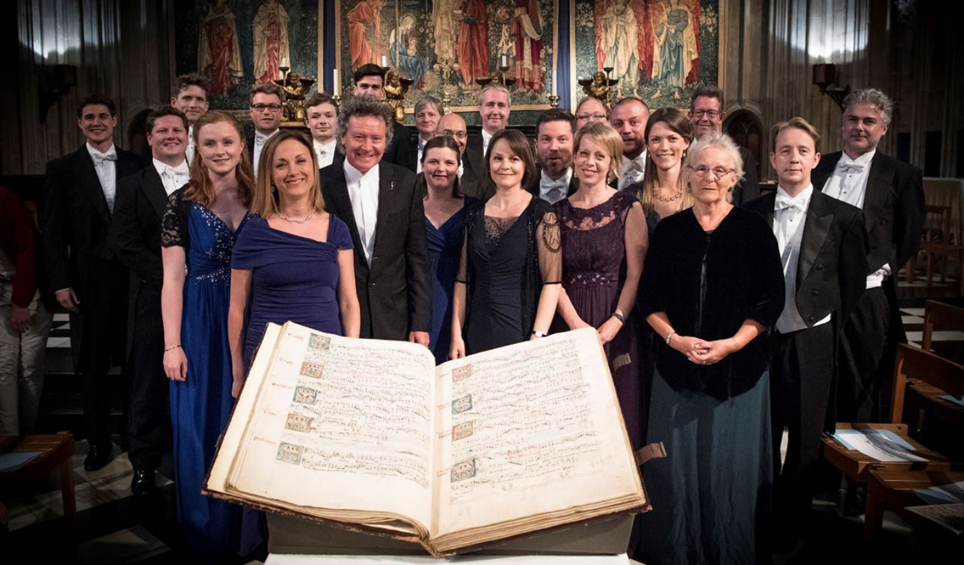 The Eton choirbook is in the centre of the image, propped open. Behind it stands Harry Christophers and members of The Sixteen choir, all dressed in formal attire, smiling at the camera.
