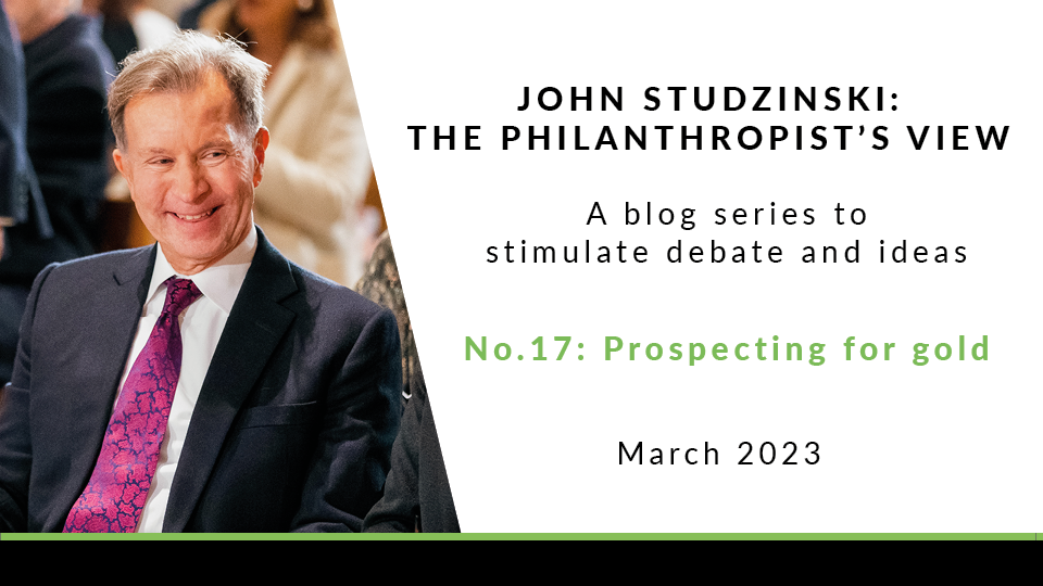 The left side of the image is a photo of John Studzinski in formal attire sat smiling amongst a busy audience. The right of the image has a white background with the text 'John Studzinski: The Philanthropist's View. A blog series. stimulate debate and ideas. No. 17. Prospecting for gold'. 