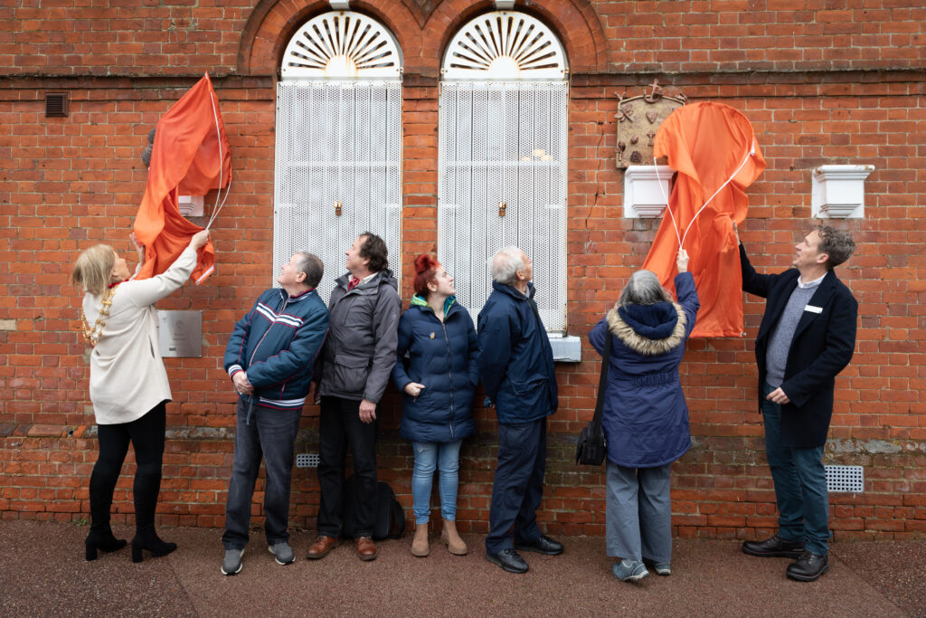 A group of people stood in front of a building with red bricks and white arched windows Either side of the group, statues beneath orange sheets are being unveiled. The group look either side of them at the statues.