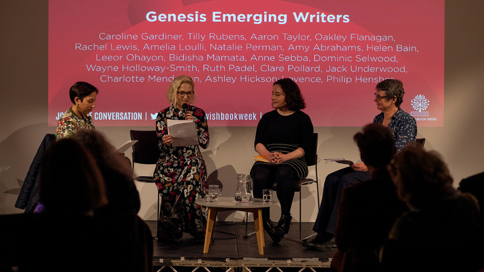 A photo of journalist Bidisha sat on a stage alongside writers Amy, Rachel and Tilly. Amy is holding up a piece of paper and speaking into a microphone The others sit and listen to her. Behind them is a projected screen with the text 'Genesis Emerging Writers' on it. In front of them is a seated audience.