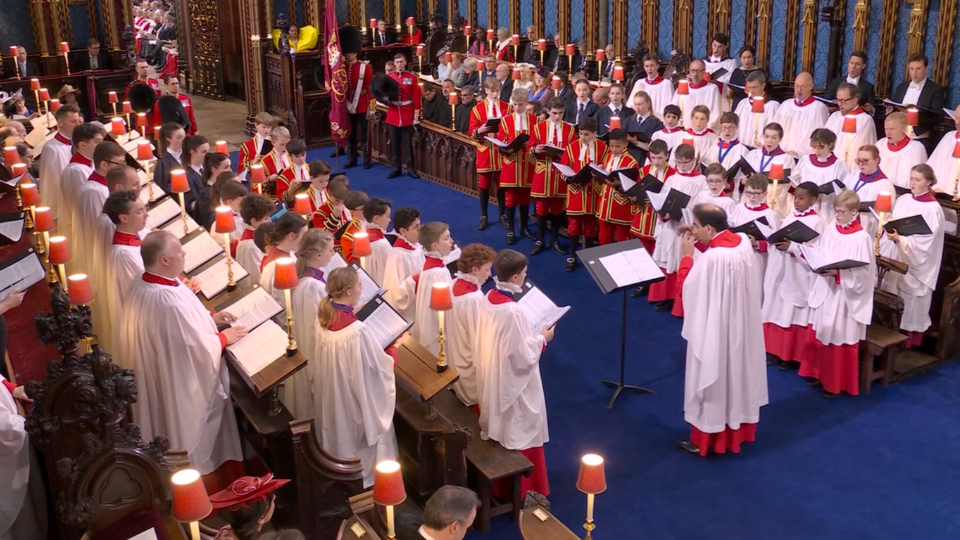 A wide shot of several rows of choral singers inside Westminster Abbey. The rows of singers are facing each other, holding scores and singing out. In the centre at one end of the singers is Andre Nethsinga, who is conducting.