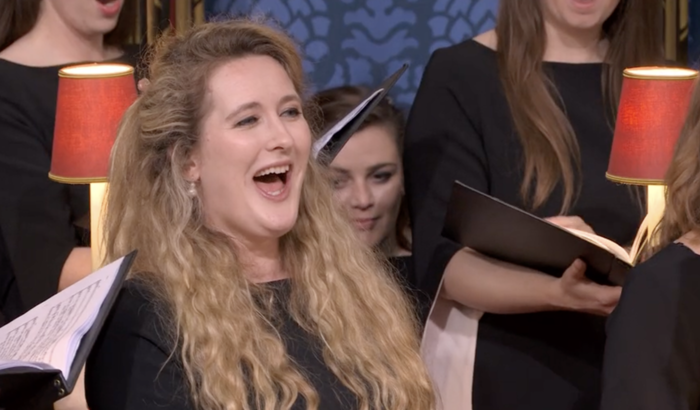 Singer Hilary Cronin stood amongst other choir members, singing out. Hilary has long blonde curly. hair and is wearing a black dress.