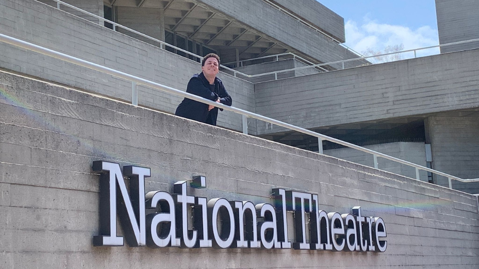 Matt Armstrong leaning against the rail of a balcony which sits above the 'National Theatre' sign on the side of the theatre building.